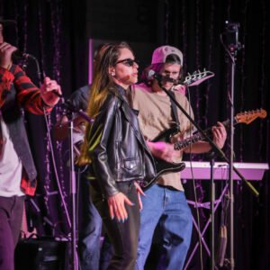 Singer in leather jacket performing on stage with live band under concert lighting.