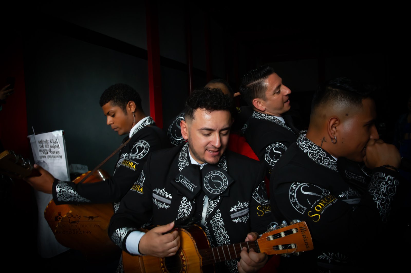 Mariachi musicians in traditional black suits playing vihuela and guitars indoors.