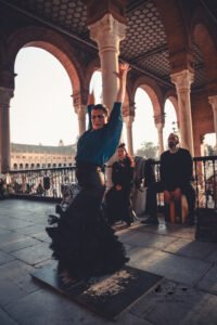 Flamenco dancer in flowing black skirt performing outdoors under arched columns.