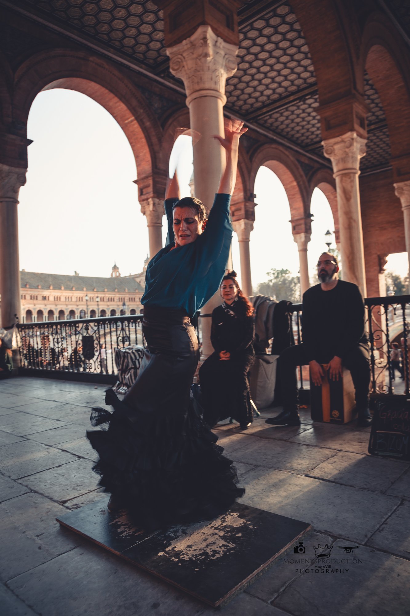 Flamenco dancer in flowing black skirt performing outdoors under arched columns.