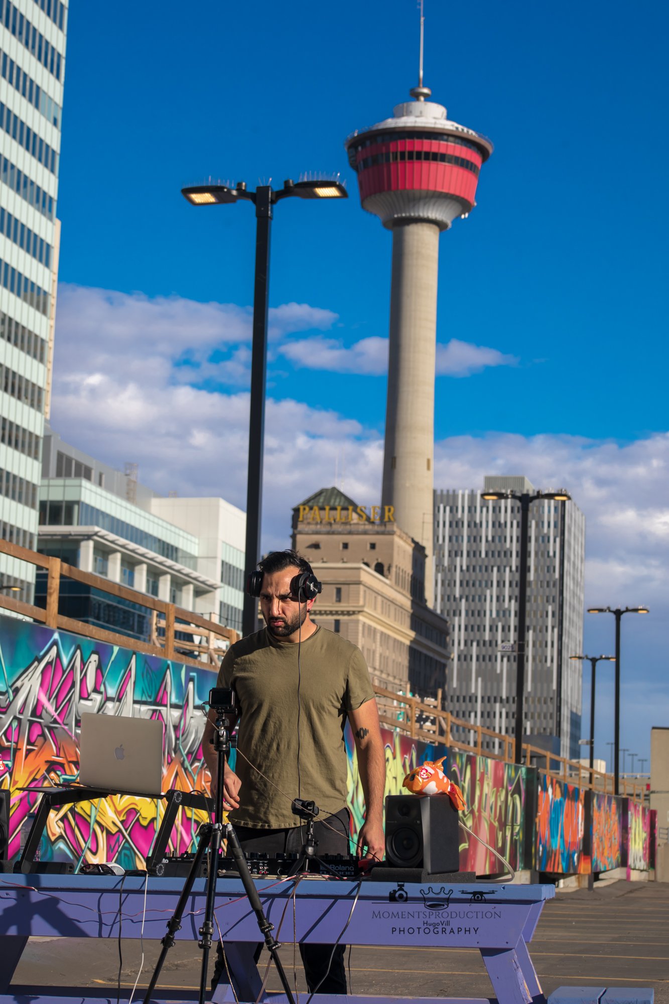 DJ performing on rooftop with mixing equipment, speakers, and urban graffiti mural behind.
